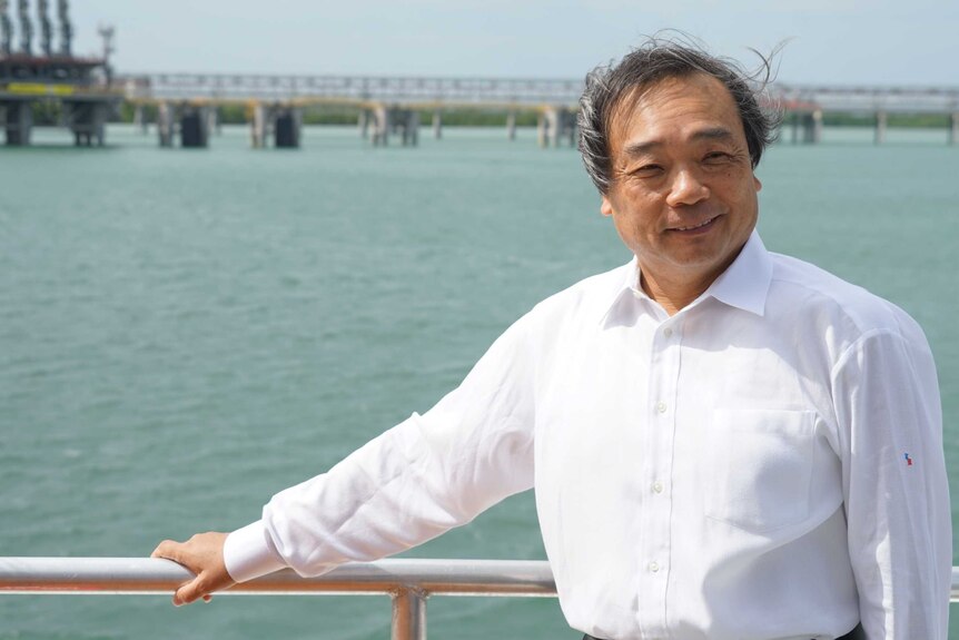 A smiling, middle-aged Japanese man, his short hair blowing in the wind as he stands in front of a harbour.