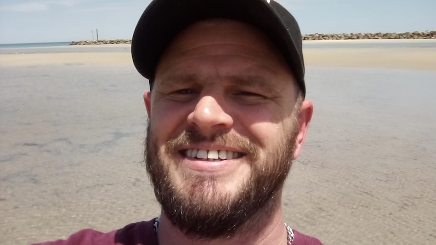 A man in a cap smiling with a beach behind him