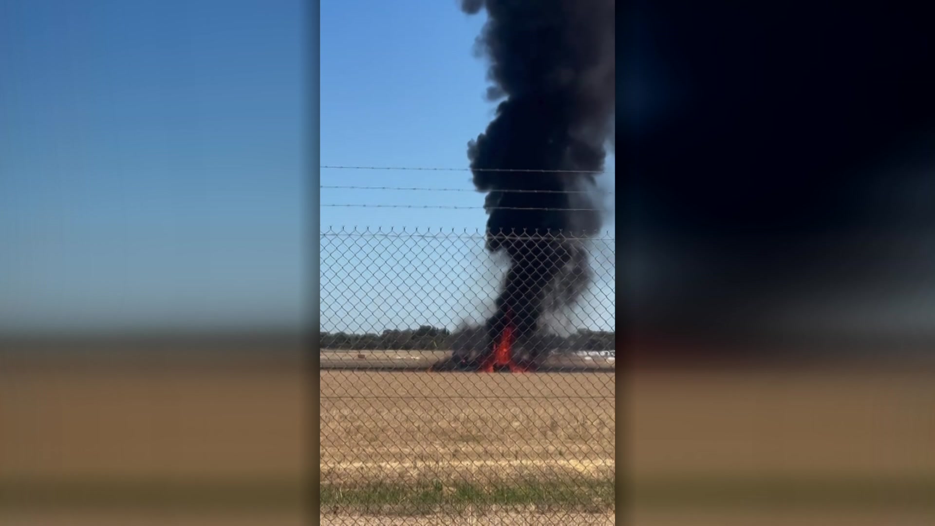 Black smoke plumes from an aircraft wreckage on an airport runway.