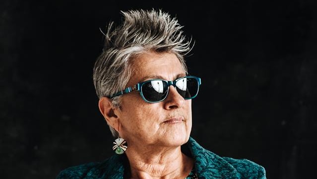 Woman with spiky grey hair, sunglasses, teal jacket, hands clasped and looking off to the distance.