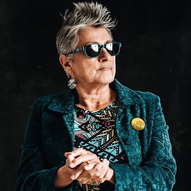 Woman with spiky grey hair, sunglasses, teal jacket, hands clasped and looking off to the distance.