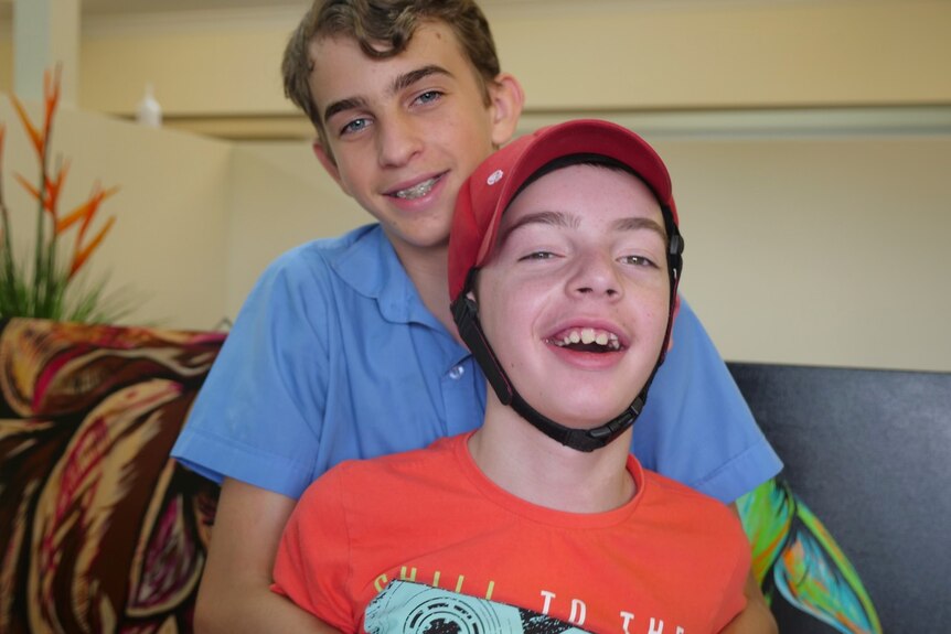 A boy, wearing a blue shirt, arms wrapped around his brother, smiling.