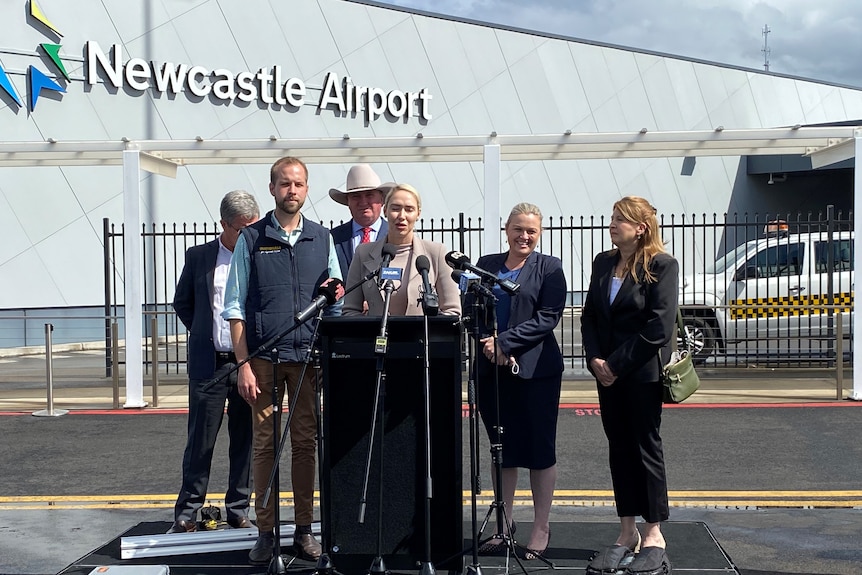 Several politicians and minders stand on a podium on a tarmac