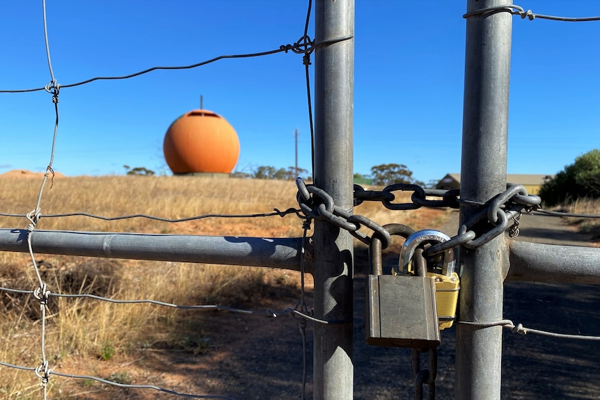 A fence with a padlock and a large orange sphere and blue sky in the background.