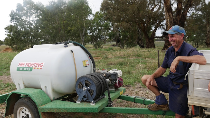 A farmer leaning on the back of his ute which is towing a small water tank. 