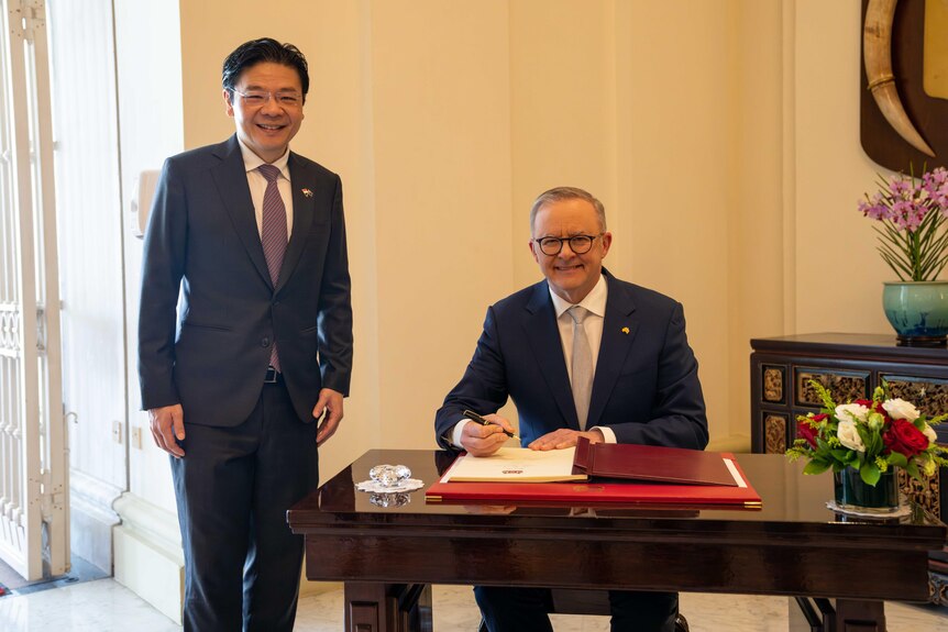 Anthony Albanese and Lawrence Wong wear suits and smile as they pose for a photo while Mr Albanese signs a document at a table.