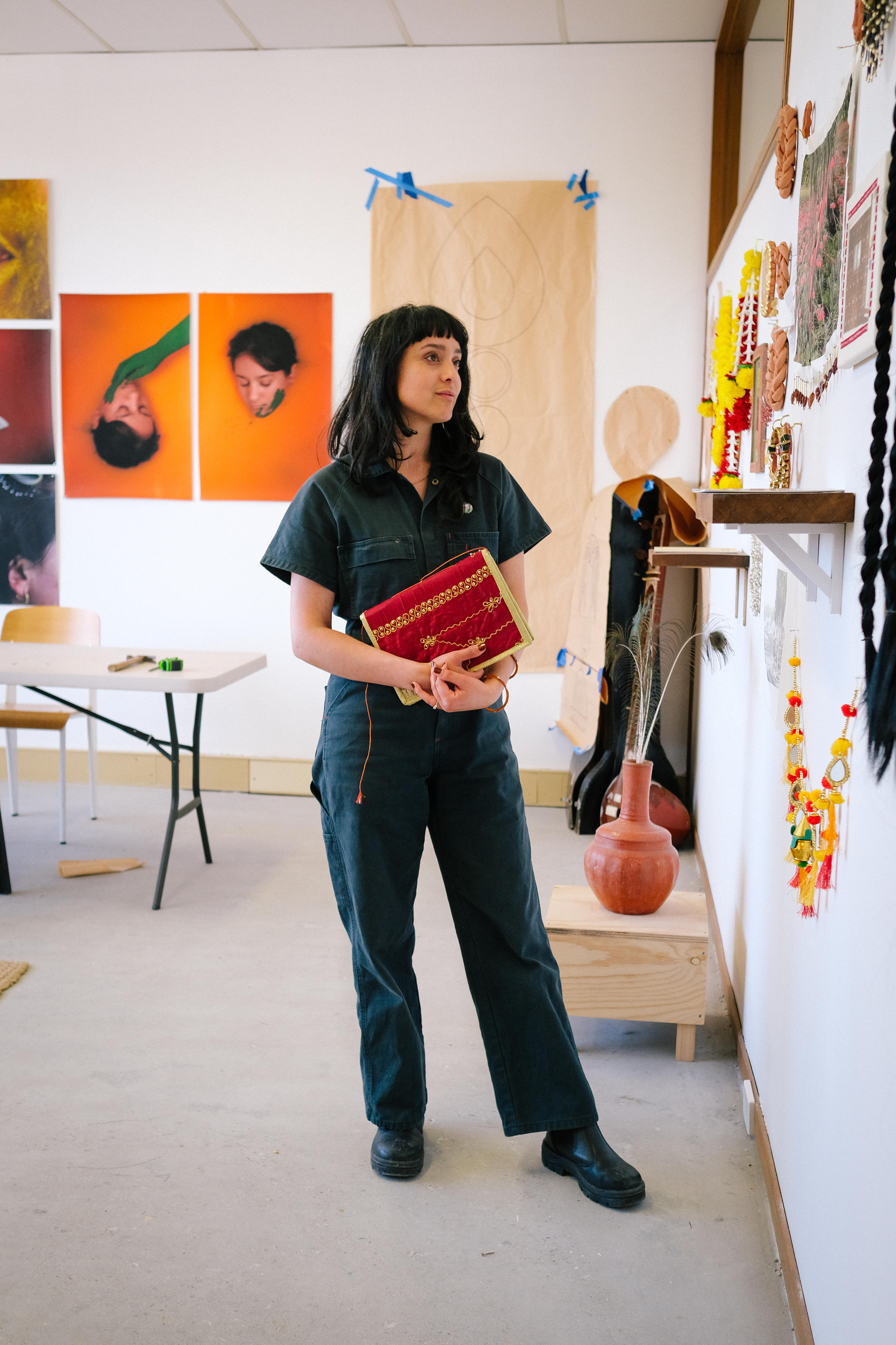 Artist Monica Rani Rudhar stands wearing a navy boiler suit and holding a red book in her studio, gazing at images on the wall. 