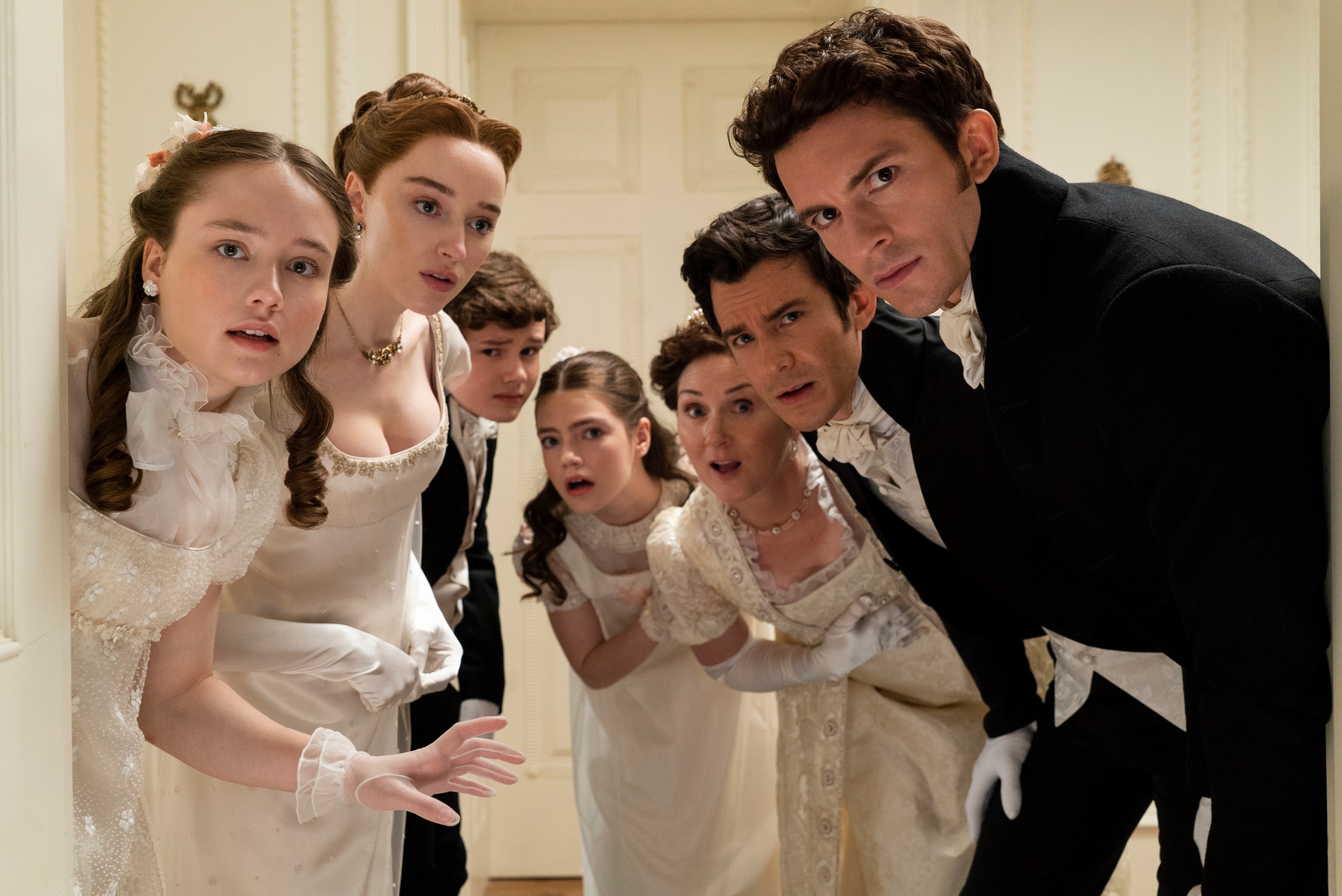 A family all wearing formal period clothing peering out of a doorway towards the camera looking surprised and alarmed