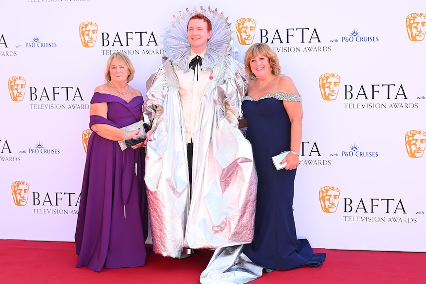 Joe Lycett, dressed in a bright silver coat and ruff, is on the red carpet flanked by two middle aged women in pretty dresses
