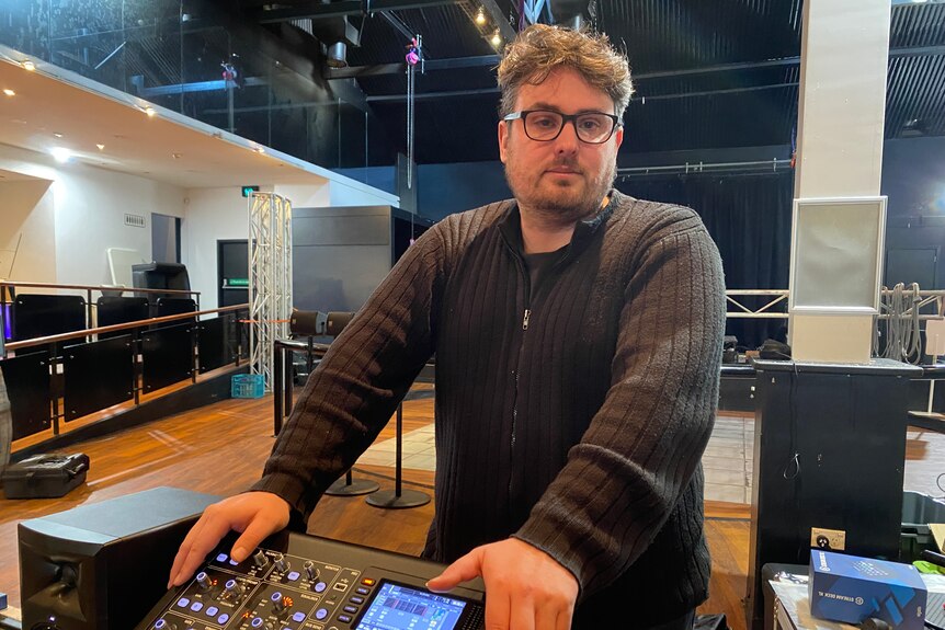 A man standing next to a sound desk with a stage in the background.