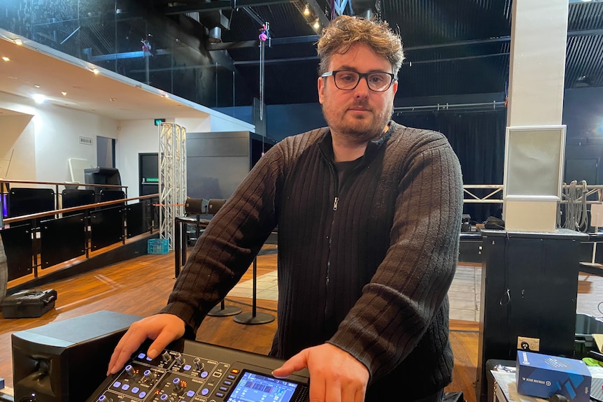 A man standing next to a sound desk with a stage in the background.