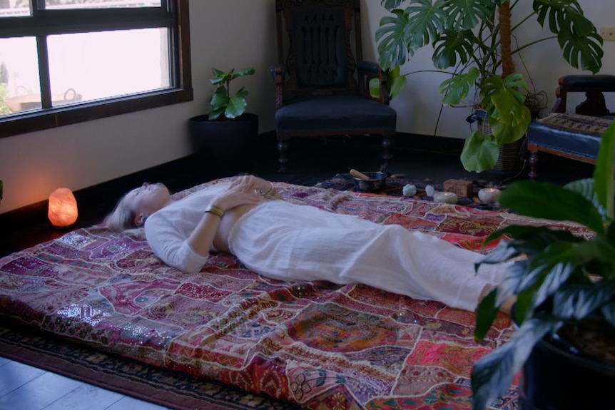 A woman in white lies on a mattress on the ground surrounded by plants. 