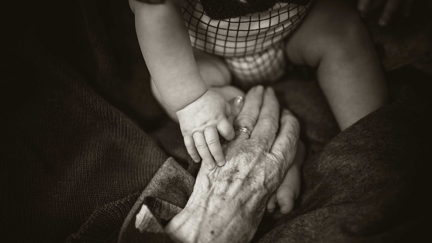A baby's hand reaches out to grab onto an older person's wrinkled fingers
