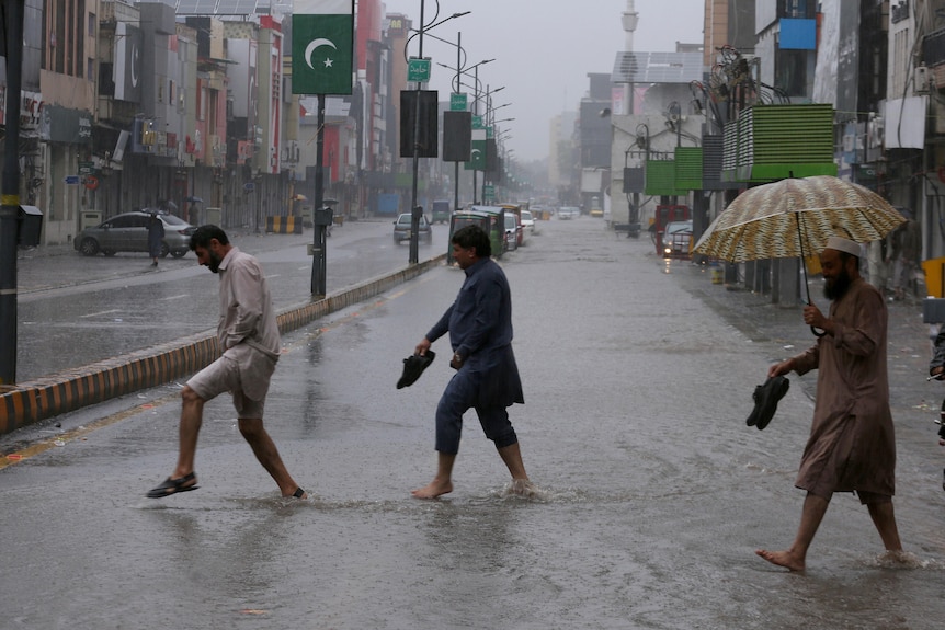Three men cross a flooded street with two holding shoes in their hands as rain tumbles down around them