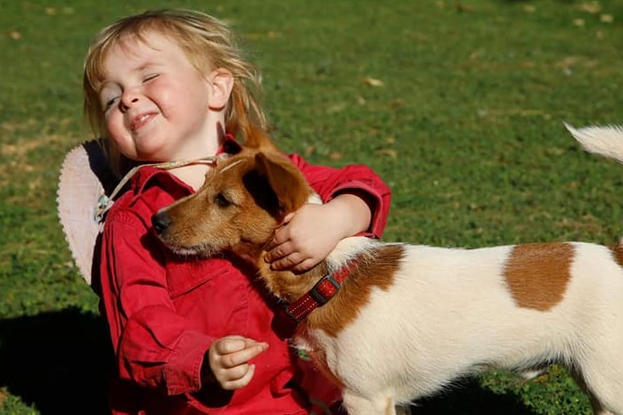 A girl in a red jacket sits on the grass hugging a small white and brown dog.
