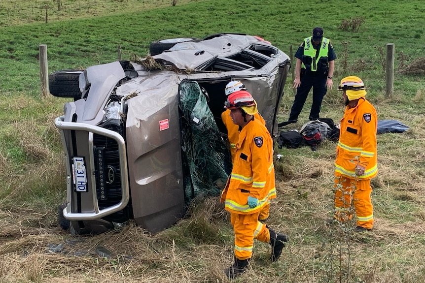 three men in hi vis gear stand next to a car rolled over on its side with a police officer standing nearby