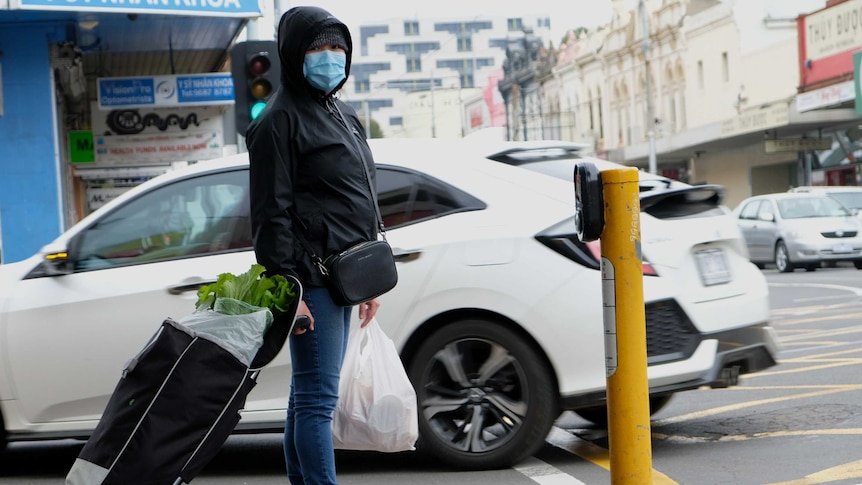 A woman wearing a mask and holding a shopping trolley waits at the lights in Footscray.