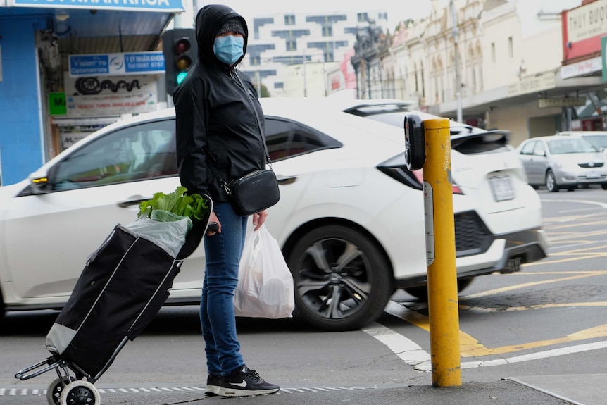 A woman wearing a mask and holding a shopping trolley waits at the lights in Footscray.