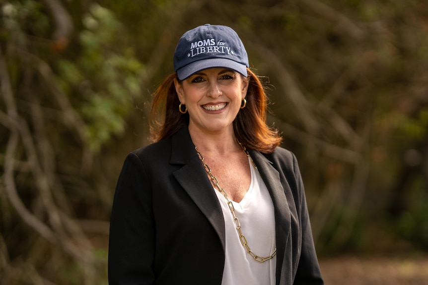 A woman in a black suit jacket and gold necklace wears a hat that says "moms for liberty"