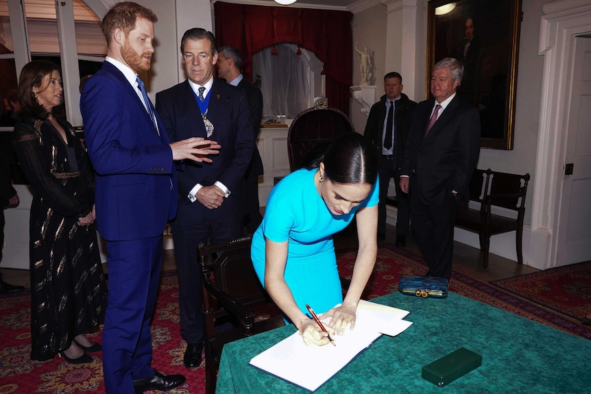 Meghan leans forward to write in a  book as Prince Harry stands behind.