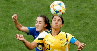 A lofted football comes towards Sam Kerr, who challenges a Brazilian player who has closed her eyes.