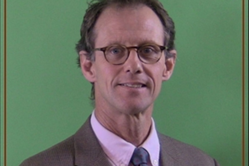 A man, wearing glasses and a suit, against a green background.