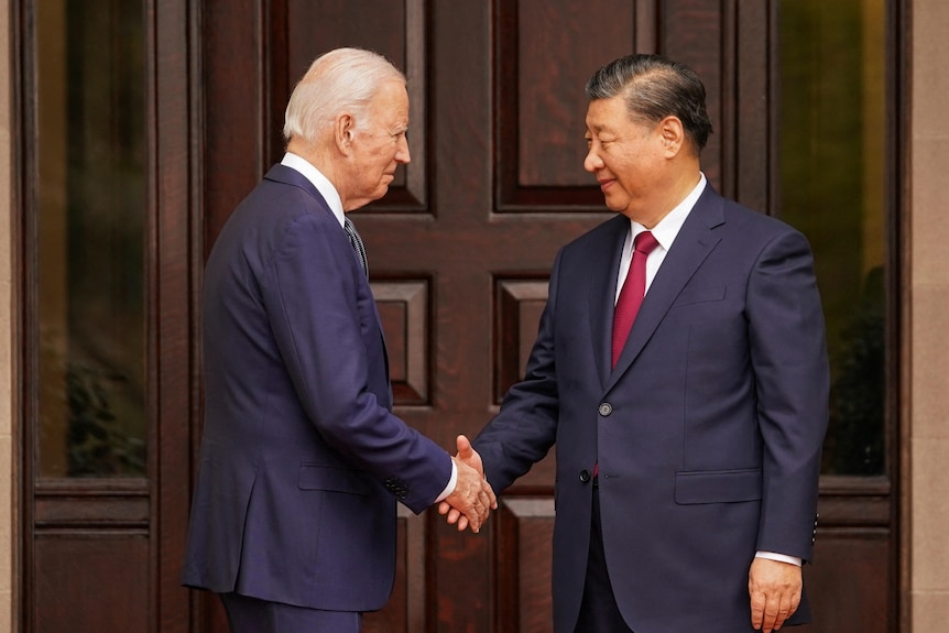 Standing in front of a large brown double door, Biden and Xi shake hands.