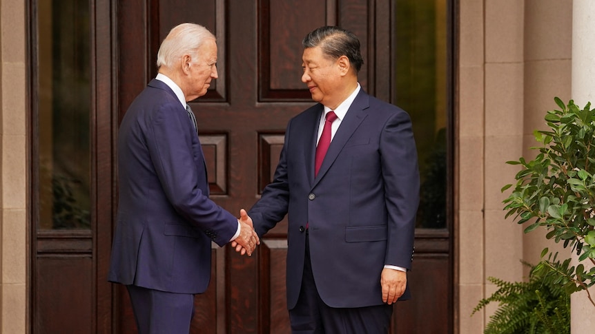 Standing in front of a large brown double door, Biden and Xi shake hands.
