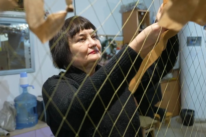 A woman with black hair weaves camouflage onto a net.