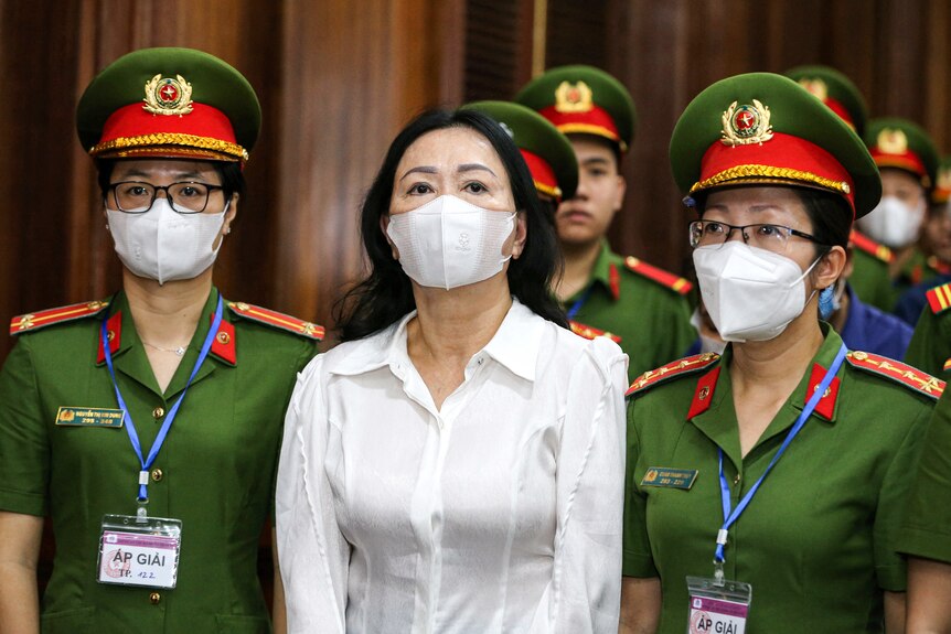 A woman wearing a white shirt and white medical mask flanked by two police women wearing green and hats