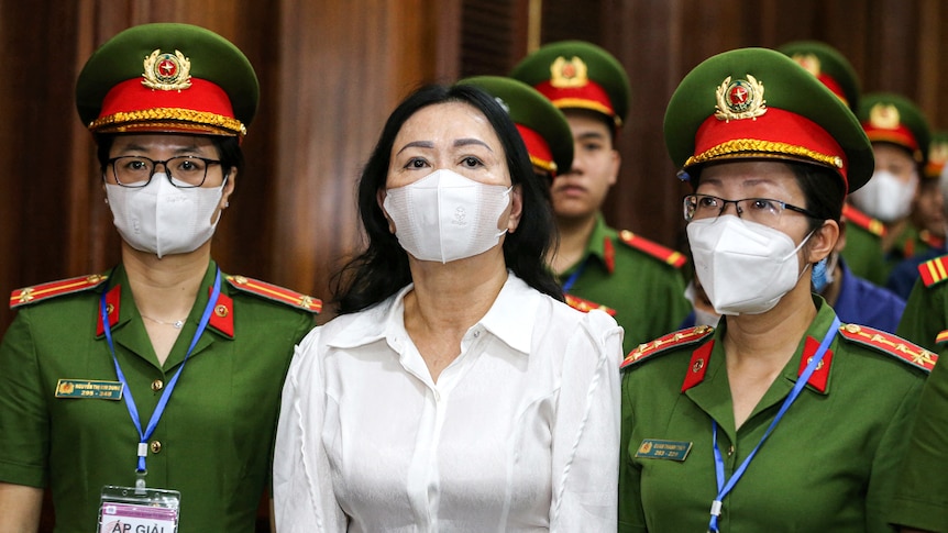 A woman wearing a white shirt and white medical mask flanked by two police women wearing green and hats