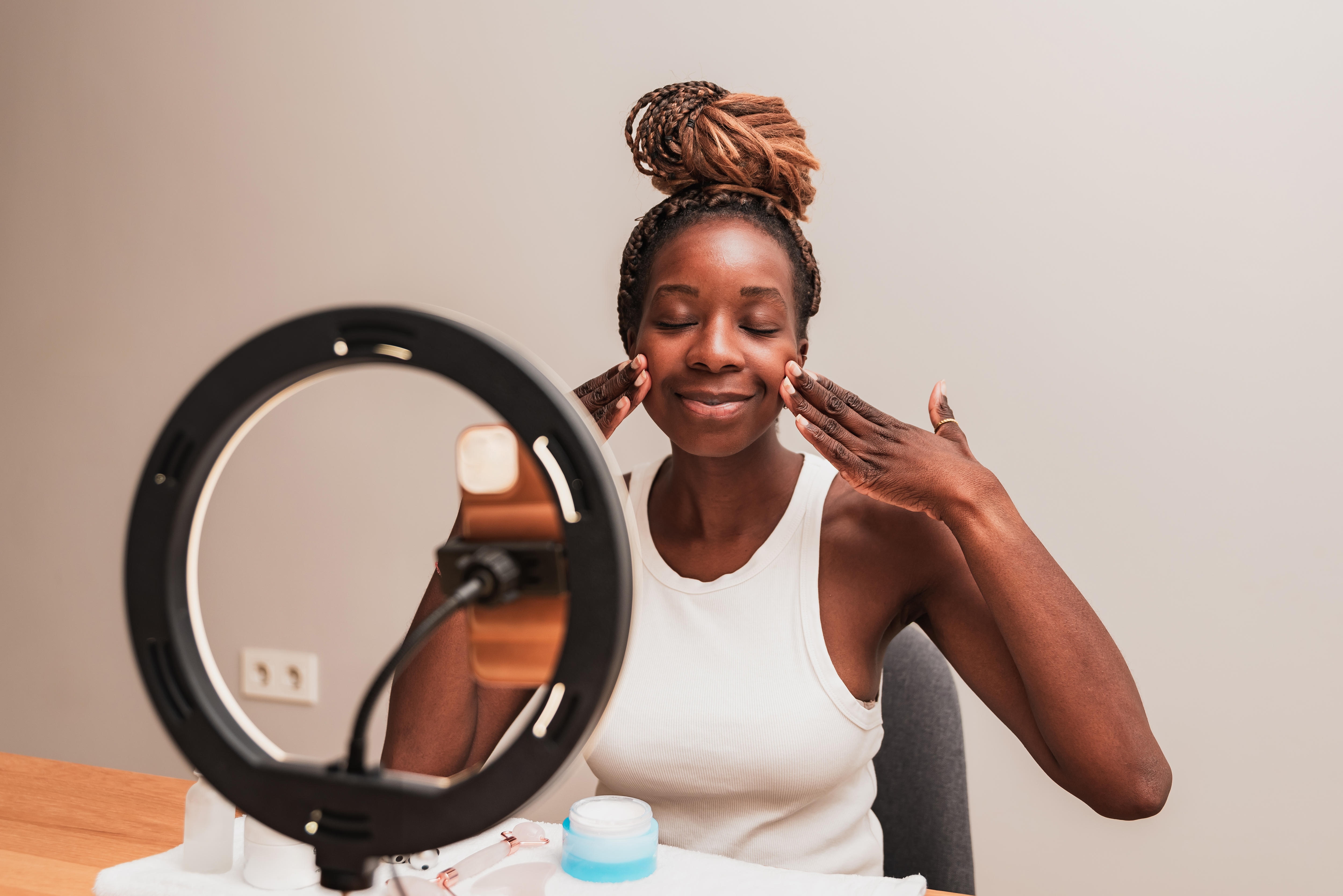 A young woman applies cream to her cheeks and smiles in front of a ring light.