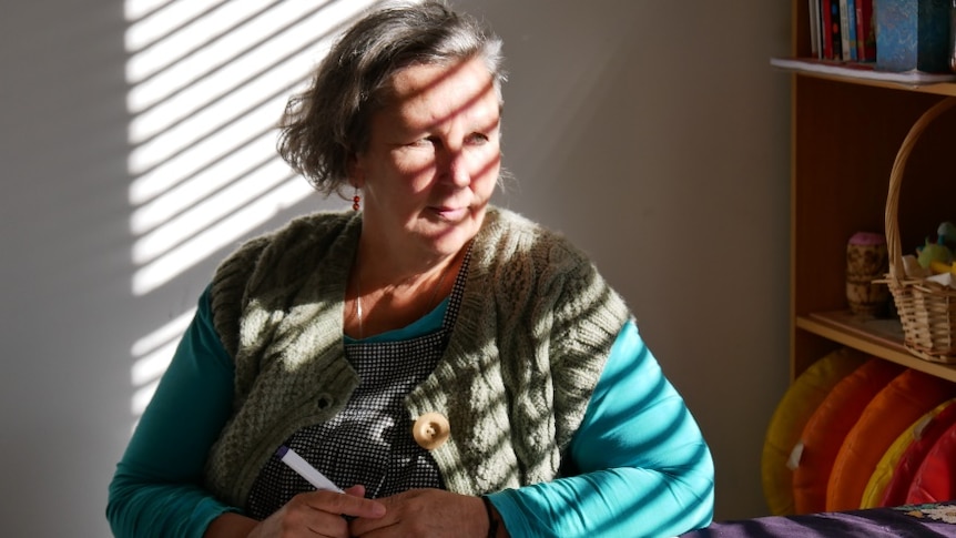 a women sits at a desk with a market in hand, wearing a blue shirt and woolen vest.