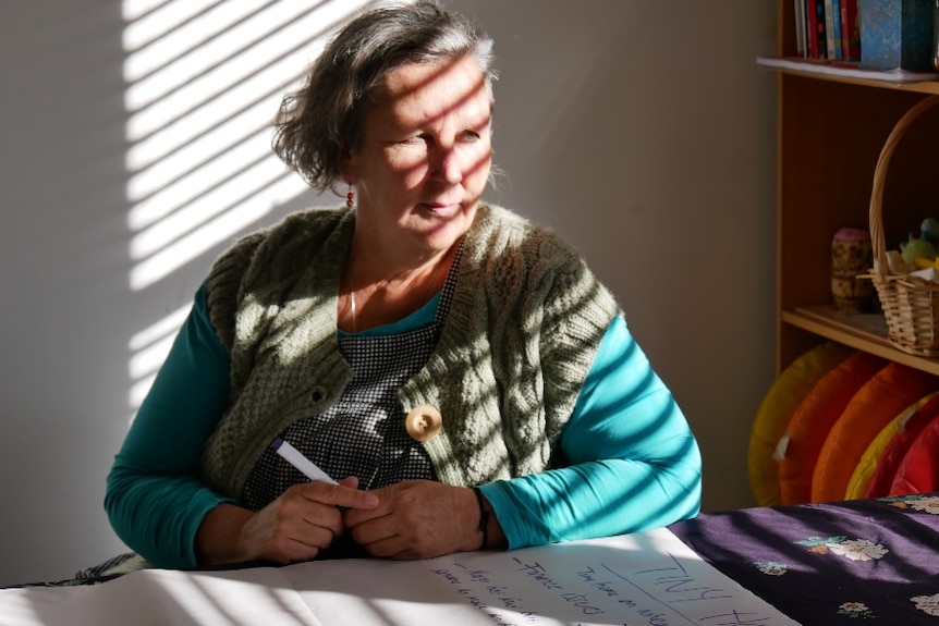 a women sits at a desk with a market in hand, wearing a blue shirt and woolen vest.