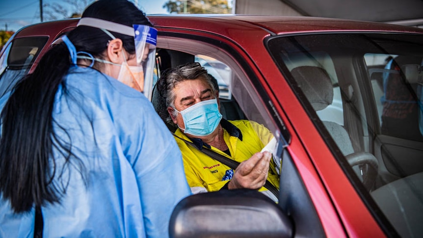 A woman wearing a medical gown and face talks with a driver through his car window. 