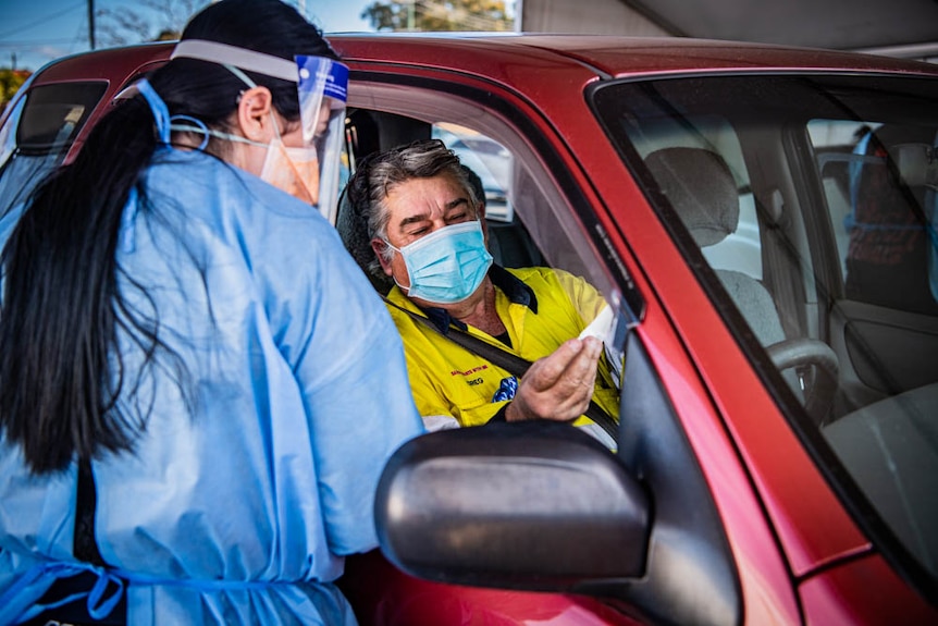 A woman wearing a medical gown and face talks with a driver through his car window. 