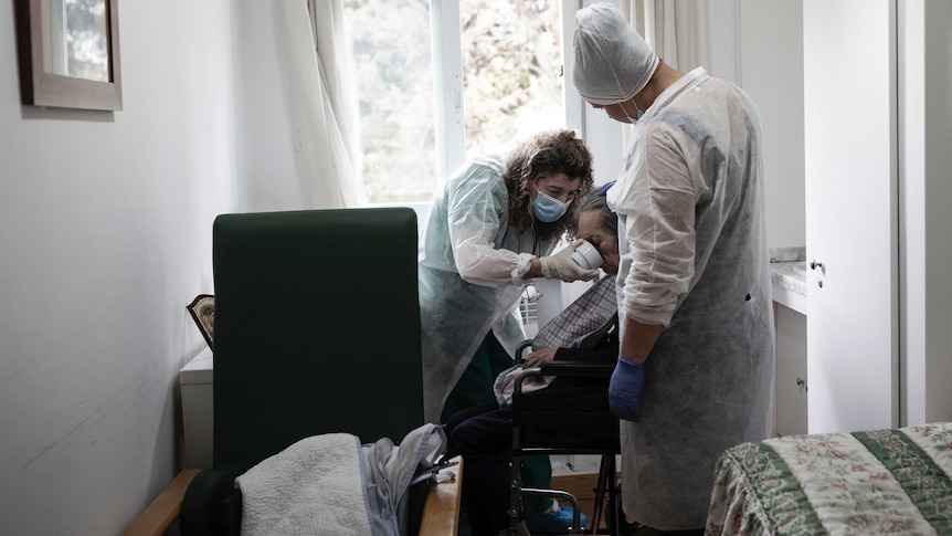 A person in a wheelchair is assisted to drink from a cup by two carers wearing masks and protective equipment.