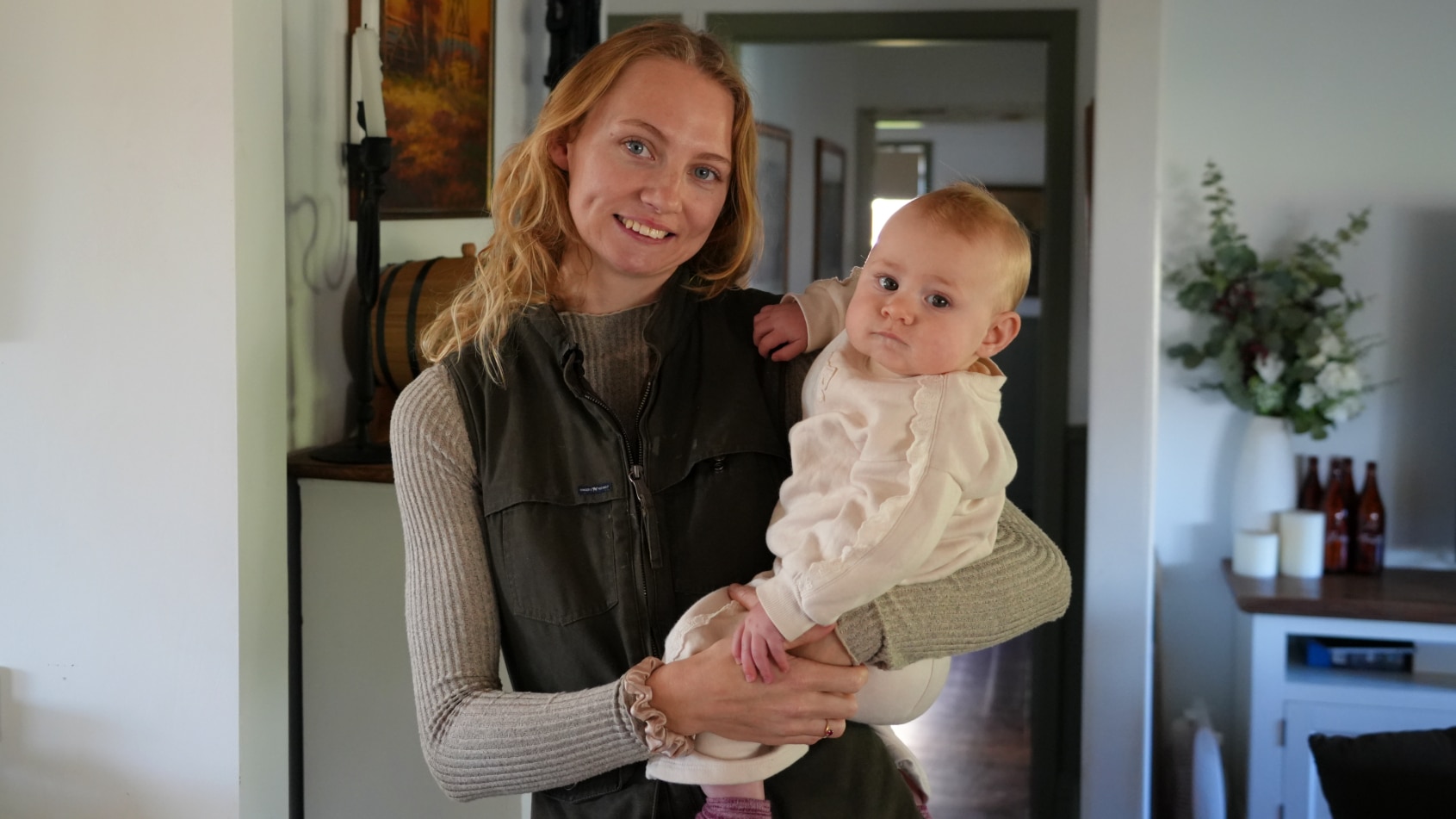 A blonde woman in a dark fleece vest and light long sleeve top looking at camera holding a baby wearing white.