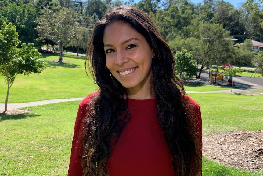 A woman with long dark wavy hair stands in a park, against the backdrop of trees and a playground.