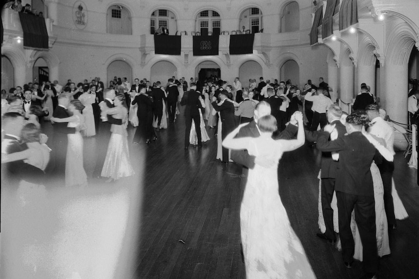 Black and white photo of circular ballroom with couples dancing arm in arm, grand pillars around the edge of room