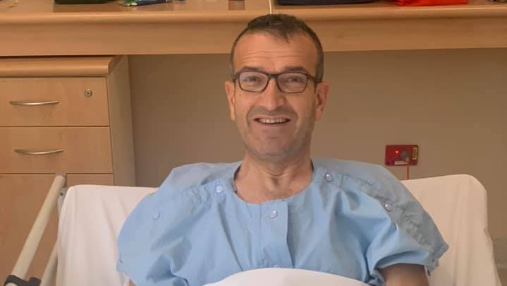 MP Tony Piccolo sits in a bed in a hospital gown, smiling at the camera