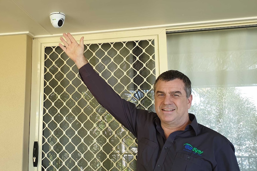 A man reaches up towards a rooftop security camera in front of a screen door