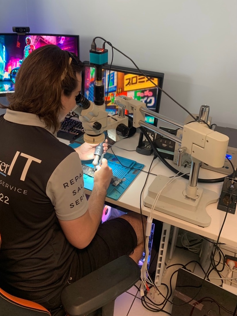 A white man with long brown hair looks into a microscope to examine a motherboard from an electronic device