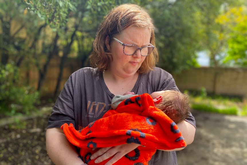 Courtney holds her son, Milo, swaddled in a red blanket as she stands in a sunlit garden.