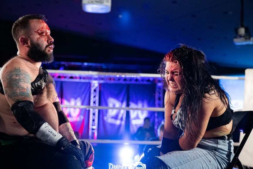 A man and woman sit facing each other in a boxing ring, both bleeding from the head and looking tired and in pain.