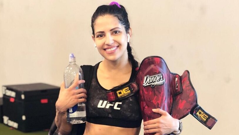 UFC fighter Polyana Viana stands in fitness gear holding a water bottle and shin guards.
