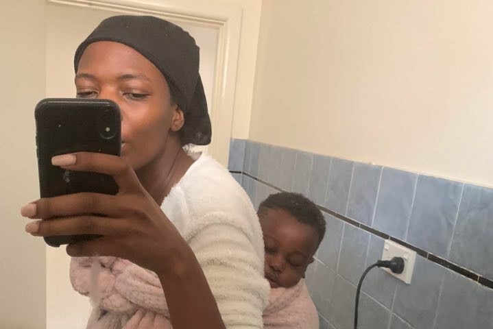 A woman takes a selfie while baby-wearing in her bathroom.
