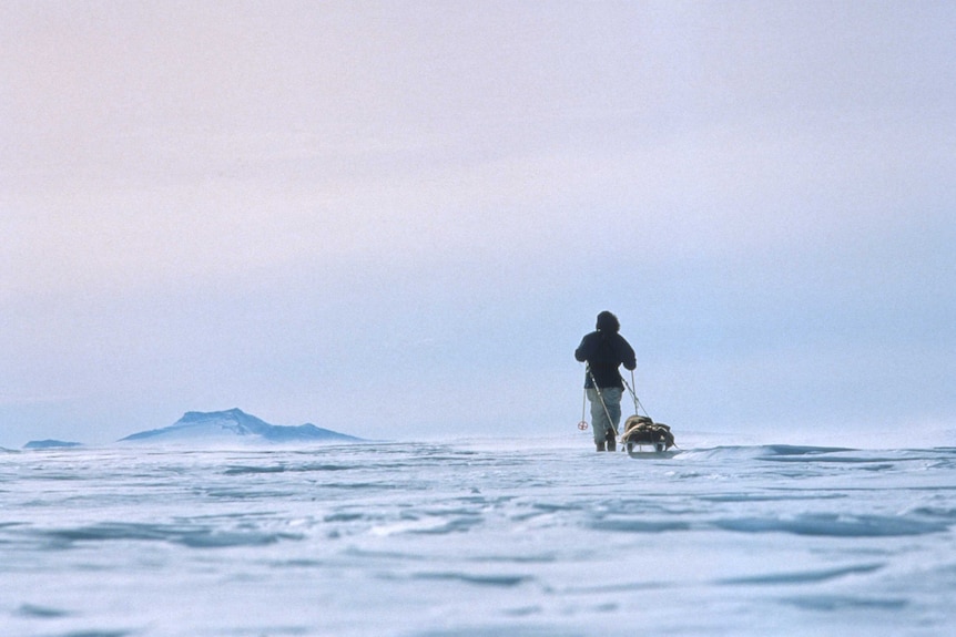 A back view of a man walking across a vast icy landscape, dragging a sled behind him.