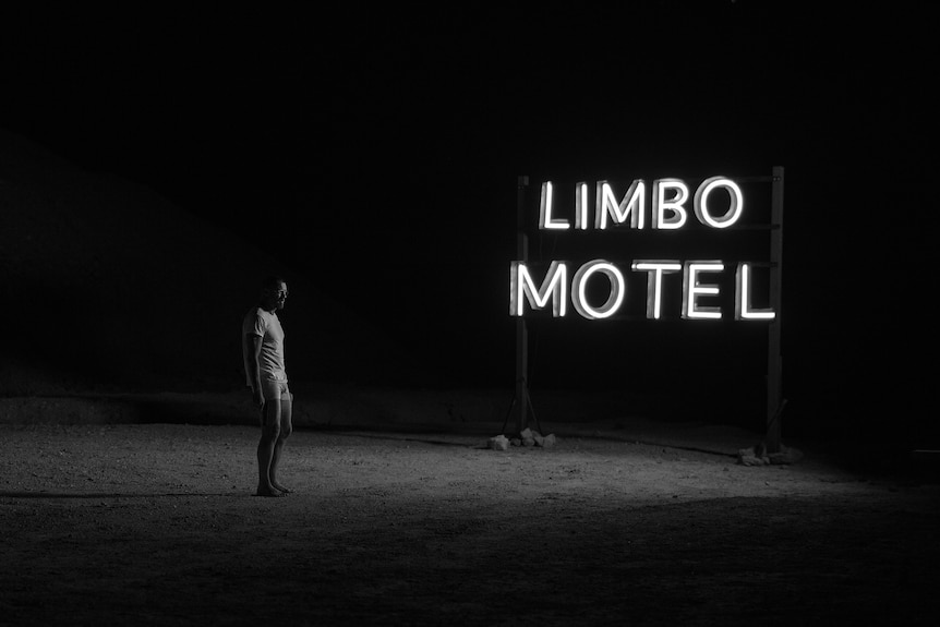 A man wearing only underpants and a t-shirt stands in the dark next to a sign that reads 'Limbo Motel'.