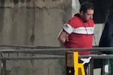 A man in a red and white shirt strides along a concrete walkway in handcuffs with a police officer beside him